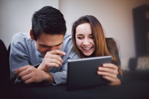 Happy young man and woman using tablet PC. Mixed race teenage couple using digital tablet smiling while lying on sofa at home.